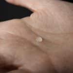 still image depicting a close up of a hand, palm up, with a single small drop of liquid in the center of the palm.