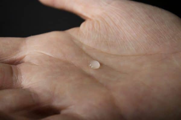 still image depicting a close up of a hand, palm up, with a single small drop of liquid in the center of the palm.