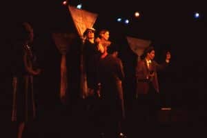 Five women on stage, dressed in formal clothing with three pieces of fabric hanging from the ceiling behind them, each tied midway. One member looks upward in distress and disappointment. One membmer looks towards the audience in shock. Another member looks towards the audience, with her arm half way up and hand in a small fist.