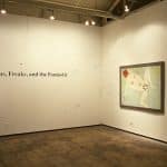 Corner of gallery, white walls with black text reading Phantoms, Freaks, and the Fantastic on the left. Right wall has two pieces of framed art. The first is a large painterly piece with a seagreen background, a white human figure laying down and leg spread, and the head is read. The next piece is small, with a red background, a white human figure with their back facing the viewer and scrunch downward.