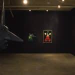 Inside a dark room, with dim lights on the pieces of painterly art on black walls. On the left is a large sculpture emerging from a mural piece of an unproportional human body doing a hand-stand. The head is creature-like with a large pointer nose, small beaded eyes, long face and neck, open mouth, and long pointed horn.