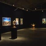 Inside a dark room, with dim lights on the pieces of painterly art on black walls. There are three sculptures displayed across the room. The two on the left are on black pedestals. The middle sculpture is a human body resembling an uncomfortable squat. The farthest sculpture is a human body from the neck to shoulders and thighs, with six rods pocking out the body.