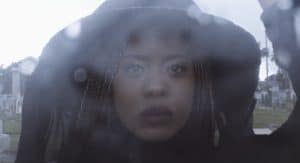 close up photograph of an African American woman with long light-colored braids, wearing a black dress, hat and veil at a cemetery. The camera lens appears to have rain drops on it.