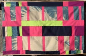 Artwork by Isela Aguirre: a cyanotype on cotton measuring 32 inches tall x 49 inches wide; the cyanotype of white feathers on blue background is pieced together like a quilt with cotton strips of vertical and horizontal rectangles of various sizes, forming an irregular grid. The cotton strips are various shades of white, pinks, bright green-yellow, magenta and black